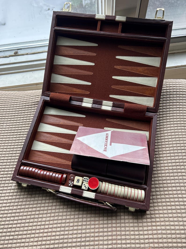 May include: Open backgammon set with a brown leather case, featuring a playing surface with alternating brown and cream-colored points. Includes dice, checkers, and a Backgammon instruction card.