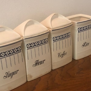 May include: Set of four white ceramic canisters with blue and white patterned designs. The canisters are labeled "Sugar", "Flour", "Coffee", and "Rice".