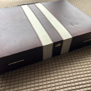 May include: A rectangular box with a burgundy leather-like exterior. Two white stripes run lengthwise across the top, separated by burgundy stripes. The stripes are stitched with white thread. The box has a small gold label.