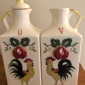 May include: A set of two white ceramic oil and vinegar bottles with rooster designs. The bottles have yellow and red accents and a floral design with a 3D rose. The bottles are labeled "O" and "V".