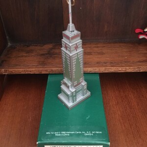 May include: A silver and green miniature ornament of a skyscraper with a gold loop for hanging. The ornament is in its original box, which is green with white text that reads "Mtd for and © 1990 Hallmark Cards, Inc. K.C. MO 64141 QK410-6 Made in China" and a barcode.