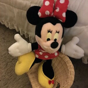 Peut inclure: Un jouet en peluche de Minnie Mouse portant un n&oelig;ud rouge &agrave; pois blancs et une robe rouge &agrave; pois blancs. Elle est assise dans un panier en osier.