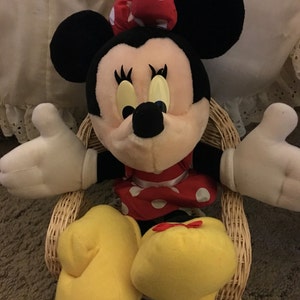 Peut inclure: Une peluche de Minnie Mouse portant une robe rouge &agrave; pois blancs et un n&oelig;ud rouge &agrave; pois blancs. Elle est assise dans un fauteuil en osier, les bras tendus.