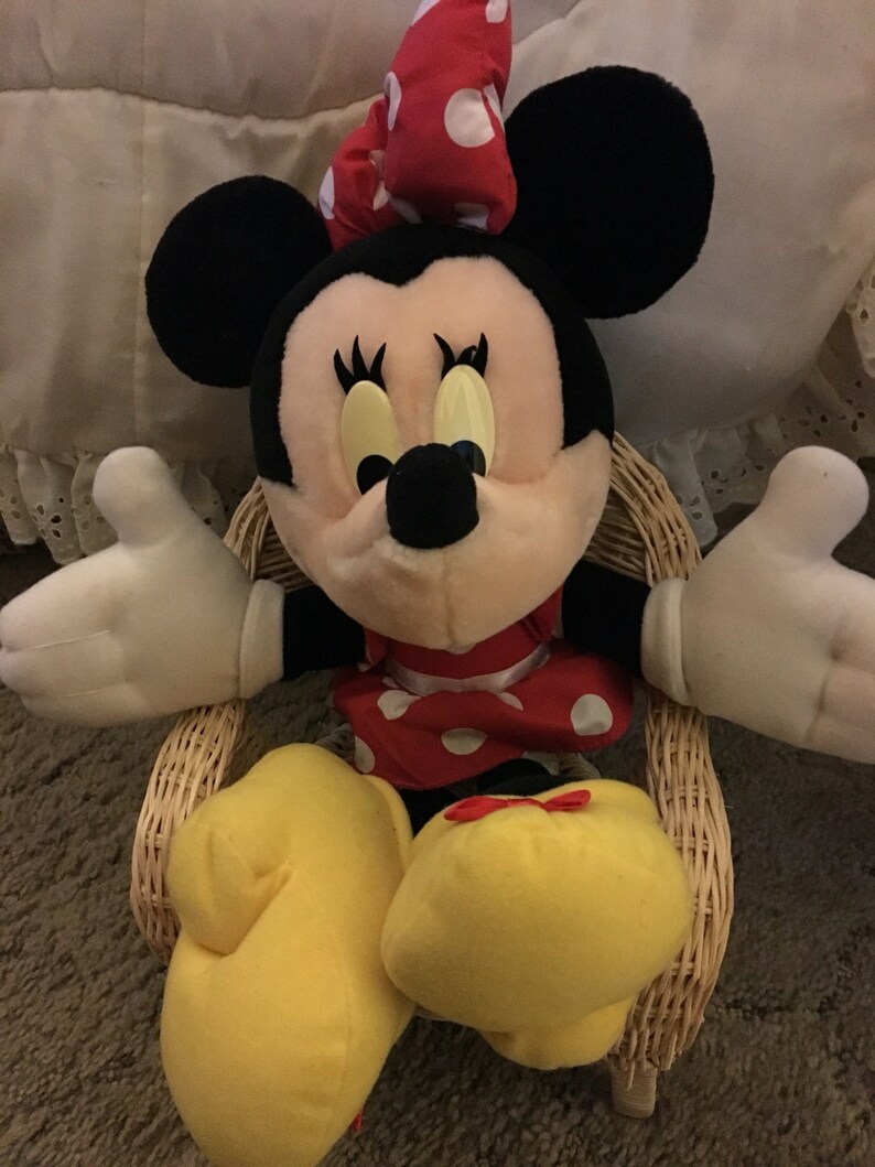 Peut inclure: Une peluche de Minnie Mouse portant une robe rouge &agrave; pois blancs et un n&oelig;ud rouge &agrave; pois blancs. Elle est assise dans un fauteuil en osier, les bras tendus.
