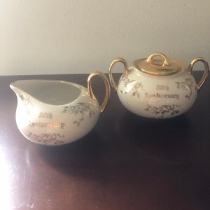 May include: A white creamer and sugar set with gold trim and floral designs. The set is decorated with the words "50th Anniversary".