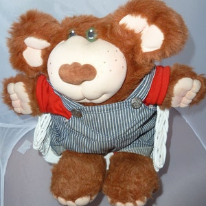 May include: A plush brown bear toy with a cream-colored face, brown nose, and green eyes. The bear wears a red shirt and blue and white striped overalls. The toy is sitting in a white wicker chair.