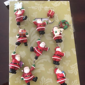 May include: A set of nine Christmas ornaments featuring Santa Claus in various poses. The ornaments are made of a red, white, and green plastic material and are strung together on a clear string. The ornaments are packaged in a box with a green and white floral pattern.