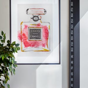 Könnte beinhalten: Ein schwarz-weiß gerahmter Druck eines Aquarells einer rosa Parfümflasche mit den Worten "COCO PARFUM CHANEL PARIS" auf dem Etikett. Ein schwarzes Lineal mit weißen Zahlen ist an der Wand neben dem Druck angebracht.
