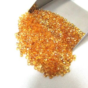 May include: A large pile of small, round, orange-brown gemstones in a metal scoop. The gemstones are loose and scattered on a white surface.