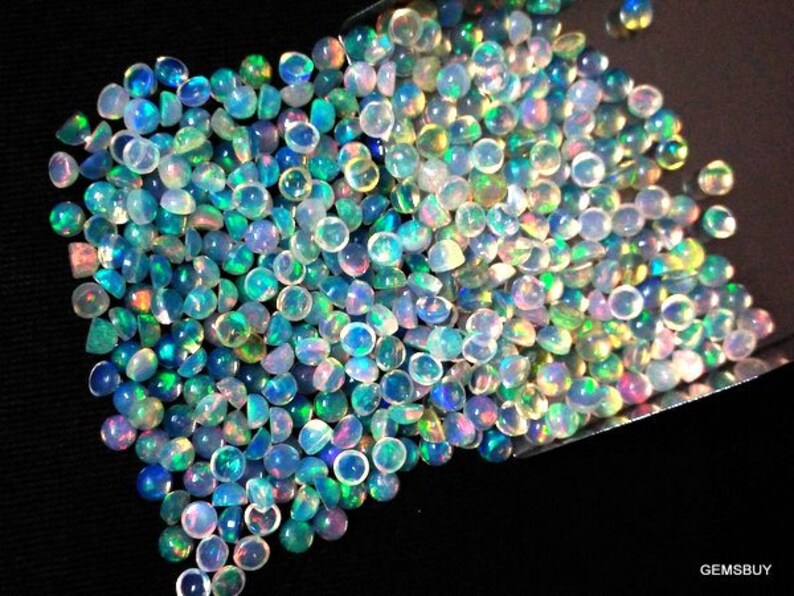 Op de afbeelding: Een stapel iriserende opaal cabochons in verschillende tinten blauw, groen en wit. De cabochons zijn rond en hebben een glad, gepolijst oppervlak.