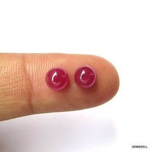 Puede incluir: Dos piedras preciosas redondas de rub&iacute; rojo, de aproximadamente 5 mm de di&aacute;metro, se muestran en un dedo.