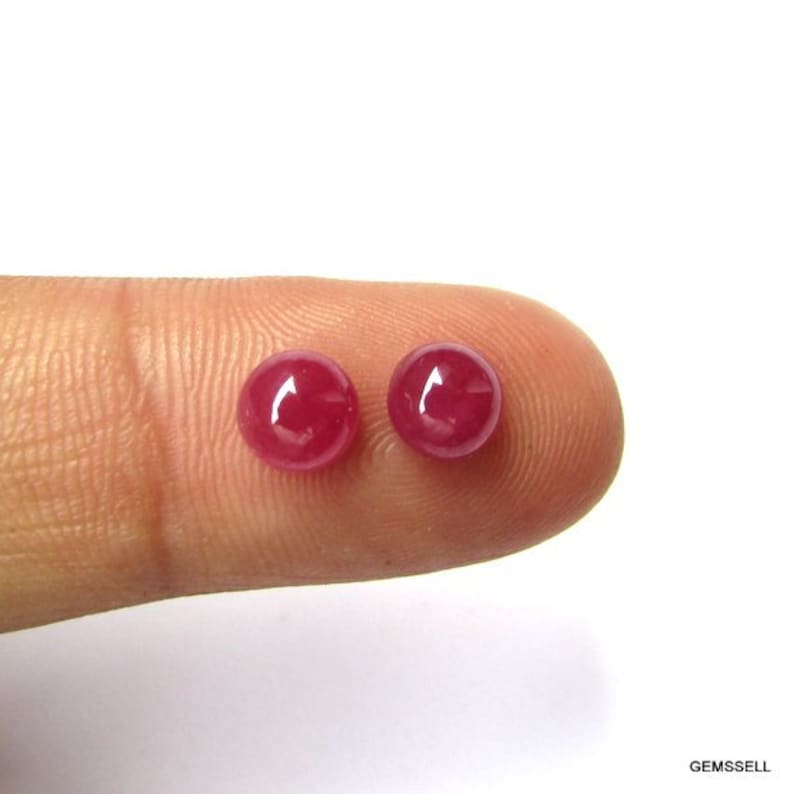 Puede incluir: Dos piedras preciosas redondas de rub&iacute; rojo, de aproximadamente 5 mm de di&aacute;metro, se muestran en un dedo.