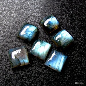 May include: Six iridescent blue and gray labradorite gemstone cabochons, cut into square shapes, displayed on a black textured surface.