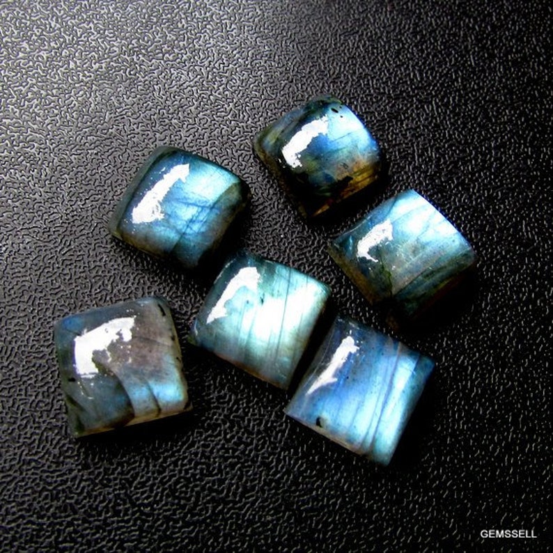 May include: Six iridescent blue and gray labradorite gemstone cabochons, cut into square shapes, displayed on a black textured surface.