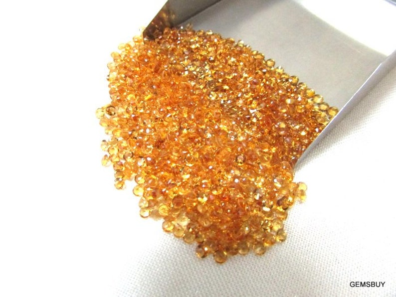 May include: A large pile of small, round, orange-brown gemstones. The gemstones are loose and scattered on a white surface.