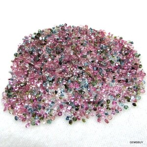 Op de afbeelding: Een grote hoop losse, gefacetteerde edelstenen in verschillende tinten roze, blauw, groen en helder. De edelstenen zijn klein en rond en verspreid over een wit oppervlak.