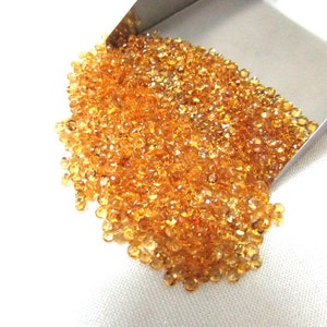May include: A large pile of small, faceted, orange-brown gemstones. The gemstones are loose and scattered on a white surface. The gemstones are spilling out of a metal scoop.