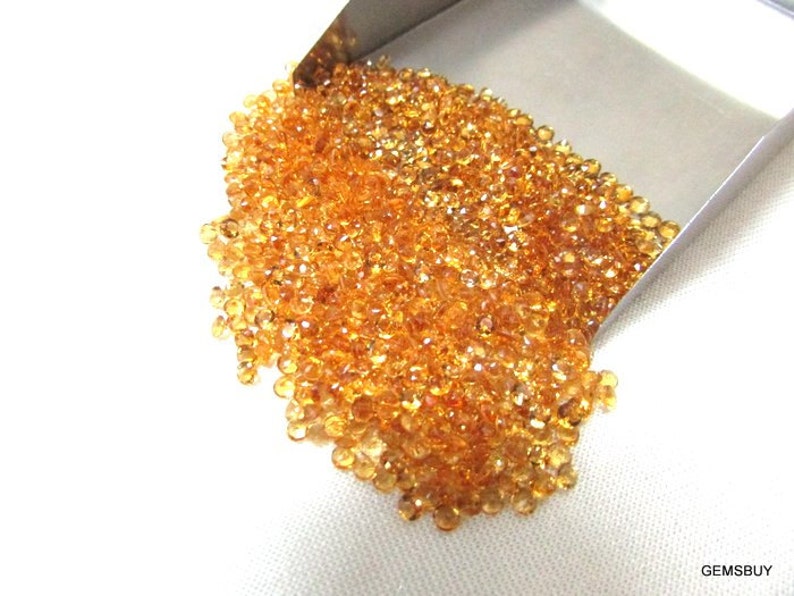 May include: A large pile of small, faceted, orange-brown gemstones. The gemstones are loose and scattered on a white surface. The gemstones are spilling out of a metal scoop.
