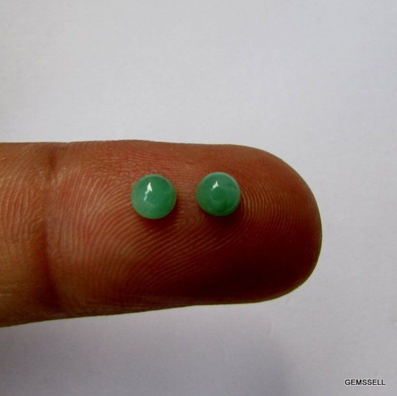 Puede incluir: Dos piedras preciosas redondas de color verde, probablemente esmeraldas, se muestran en un dedo. Las piedras preciosas son lisas y pulidas.