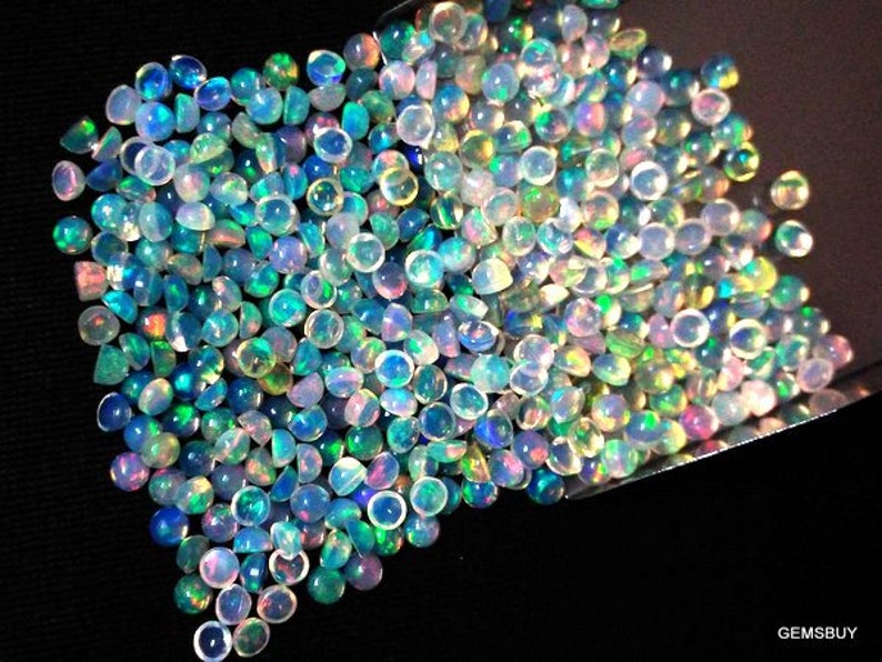 Op de afbeelding: Een stapel iriserende opaal cabochons in verschillende tinten blauw, groen en wit. De cabochons zijn rond en hebben een glad, gepolijst oppervlak.