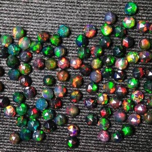 Puede incluir: Una colecci&oacute;n de opales negros sueltos, facetados, iridiscentes con destellos verdes, rojos y azules. Los opales est&aacute;n dispuestos en forma de coraz&oacute;n sobre una superficie texturizada gris oscuro.