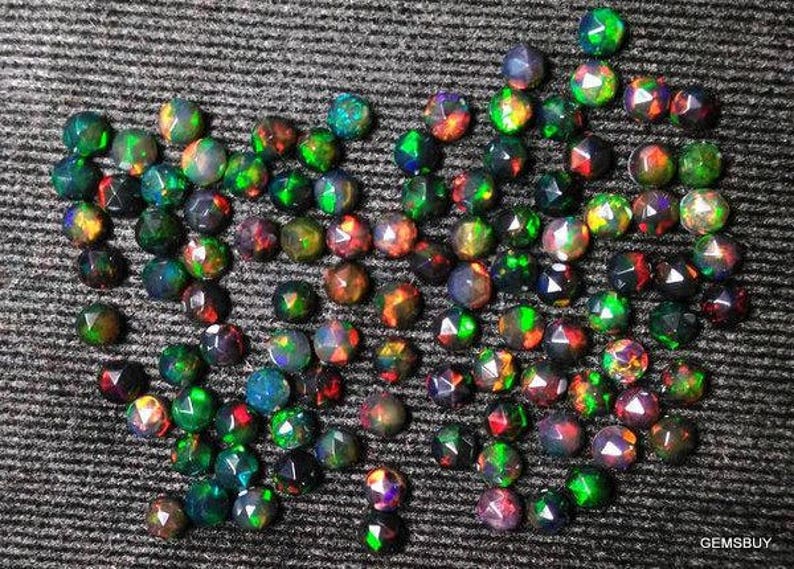 Puede incluir: Una colecci&oacute;n de opales negros sueltos, facetados, iridiscentes con destellos verdes, rojos y azules. Los opales est&aacute;n dispuestos en forma de coraz&oacute;n sobre una superficie texturizada gris oscuro.
