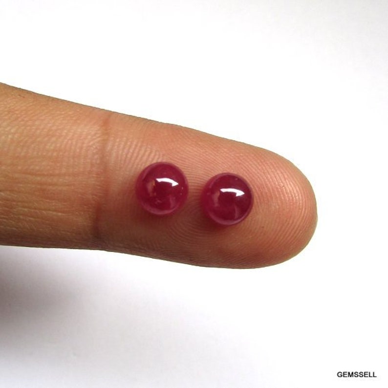 Puede incluir: Dos cabujones redondos de rub&iacute; rojo, de aproximadamente 5 mm de di&aacute;metro, se muestran en un dedo.