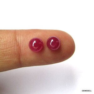 Puede incluir: Dos cabujones redondos de rub&iacute; rojo, de aproximadamente 6 mm de di&aacute;metro, se muestran en un dedo.