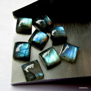 May include: A group of eight iridescent blue-green labradorite gemstone cabochons, cut into square shapes, displayed on a silver tray.
