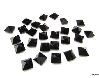 May include: A collection of 25 black square pyramid-shaped gemstones. The gemstones are arranged in a random pattern on a white surface.