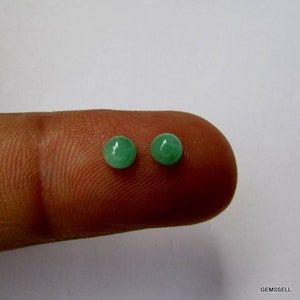 Puede incluir: Dos piedras preciosas redondas de color verde, probablemente esmeraldas, se muestran en un dedo. Las piedras preciosas son lisas y pulidas.