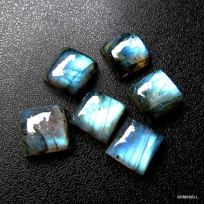 May include: Six iridescent blue-green labradorite gemstone cabochons, cut into square shapes, displayed on a black textured surface.