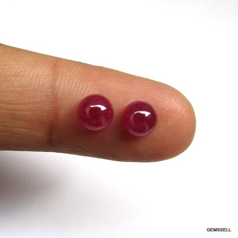 Puede incluir: Dos piedras preciosas redondas de rub&iacute; rojo, cada una de aproximadamente 5 mm de di&aacute;metro, se muestran en un dedo.