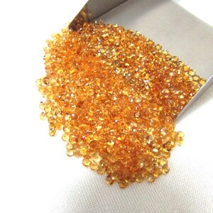 May include: A large pile of small, round, orange-brown gemstones. The gemstones are loose and scattered on a white surface.