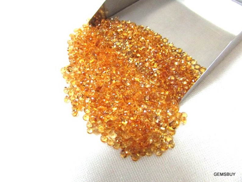 May include: A large pile of small, round, orange-brown gemstones. The gemstones are loose and scattered on a white surface.