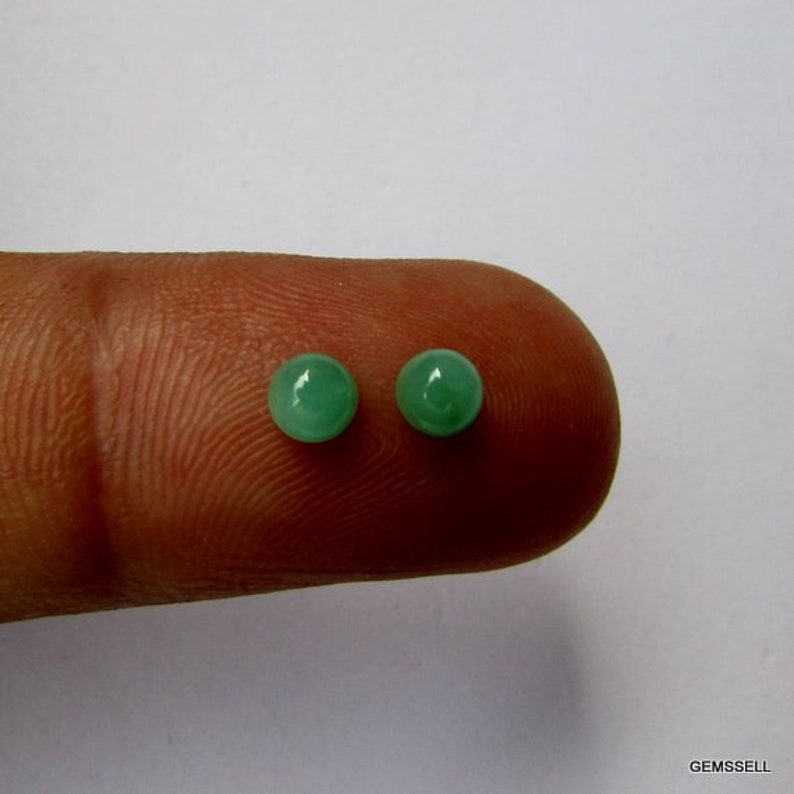 Puede incluir: Dos piedras preciosas redondas de color verde, probablemente esmeraldas, se muestran en un dedo. Las piedras preciosas son lisas y pulidas.