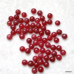 May include: A collection of round, red ruby gemstones. The gemstones are smooth and polished, and they are scattered on a white surface.