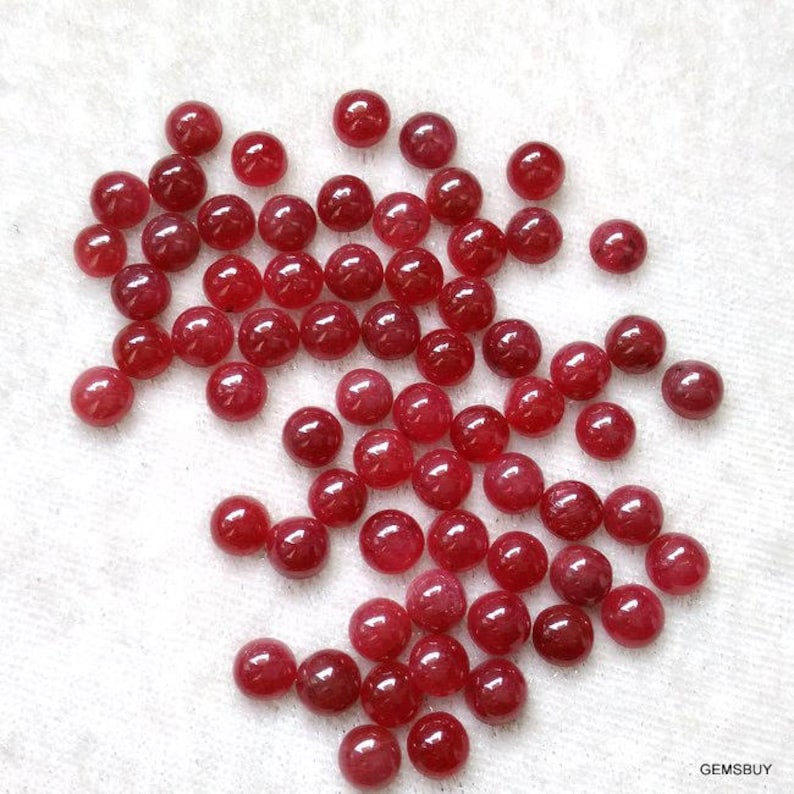 May include: A collection of round, red ruby gemstones. The gemstones are smooth and polished, and they are scattered on a white surface.