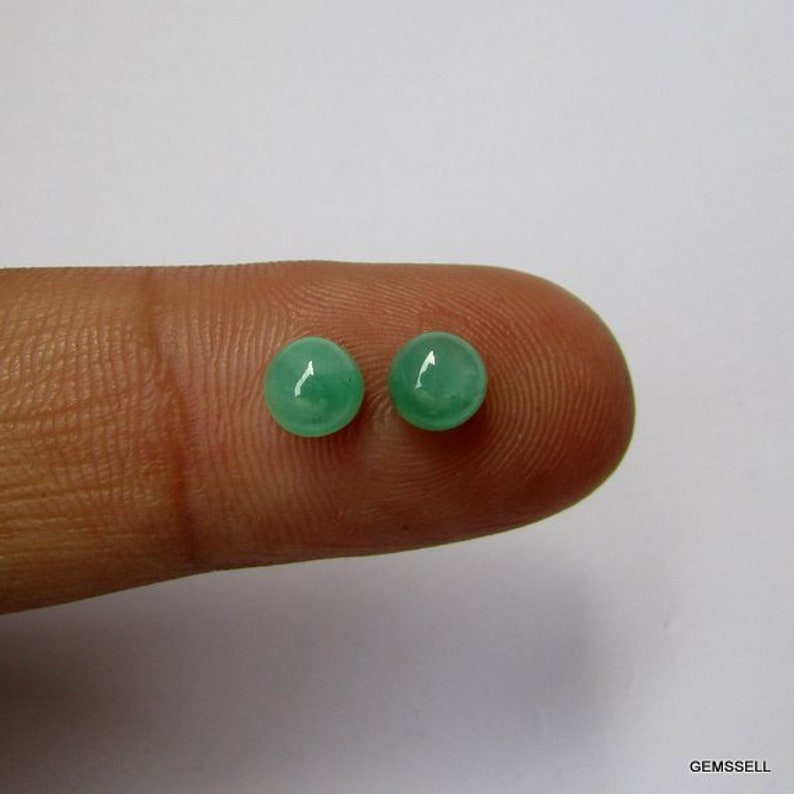 Op de afbeelding: Twee ronde, groene edelsteen cabochons, ongeveer 6 mm in diameter, worden getoond op een vinger.