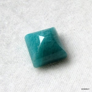 May include: A square-shaped, faceted, teal green gemstone. The gemstone has a smooth, polished surface.