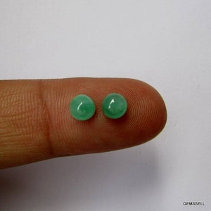 Op de afbeelding: Twee ronde, groene edelsteen cabochons, ongeveer 6 mm in diameter, worden getoond op een vinger.