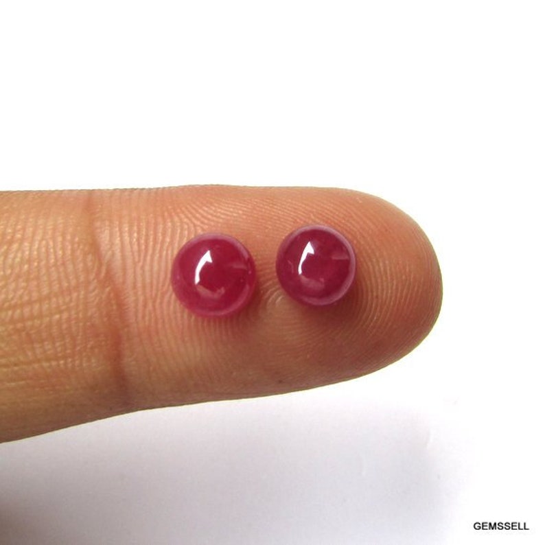 Puede incluir: Dos cabujones redondos de rub&iacute; rojo, de aproximadamente 5 mm de di&aacute;metro, se muestran en un dedo.