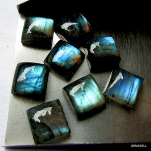 May include: A collection of eight iridescent blue-green labradorite gemstones, cut into square shapes. The stones are arranged on a silver surface.