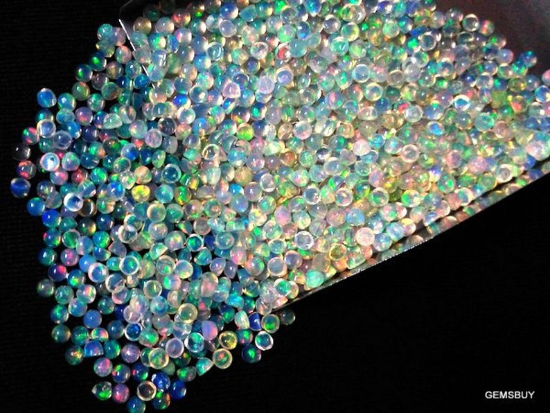 Op de afbeelding: Een grote stapel iriserende opaal cabochons in verschillende tinten blauw, groen en roze. De opalen zijn rond en hebben een gladde, gepolijste afwerking.