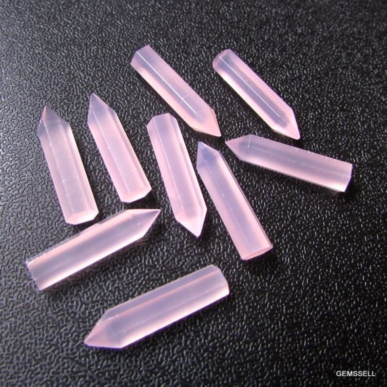 Puede incluir: Diez puntas de cristal rosa, cada una con una punta puntiaguda y una base plana, dispuestas en forma de estrella sobre una superficie negra.