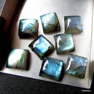 May include: Eight square-shaped labradorite gemstones with iridescent blue and green hues. The stones are arranged in a cluster on a silver surface.