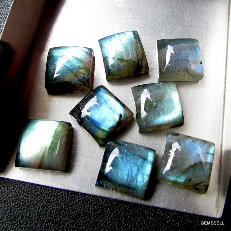 May include: Eight square-shaped labradorite gemstones with iridescent blue and green hues. The stones are arranged in a cluster on a silver surface.