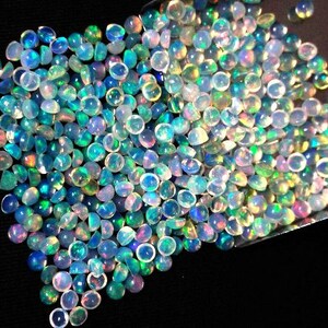Op de afbeelding: Een grote stapel iriserende opaal cabochons in verschillende tinten blauw, groen en wit. De opalen zijn rond en hebben een gladde, gepolijste afwerking.