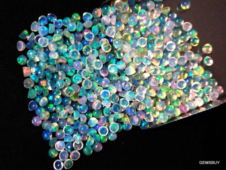 Op de afbeelding: Een grote stapel iriserende opaal cabochons in verschillende tinten blauw, groen en wit. De opalen zijn rond en hebben een gladde, gepolijste afwerking.