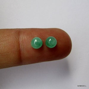 Op de afbeelding: Twee ronde, groene edelstenen, waarschijnlijk smaragden, worden op een vinger getoond. De edelstenen zijn glad en gepolijst.